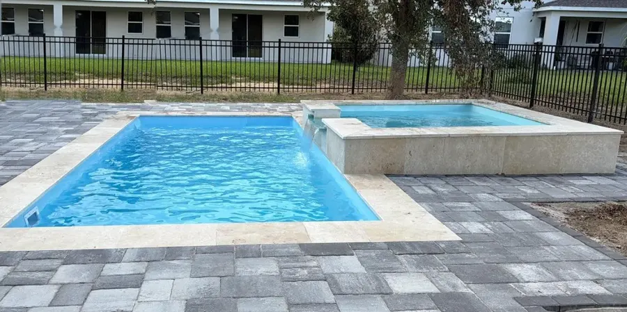 Piscina de fibra de vidrio Homery con spa elevado con desborde, coping de travertino y deck de pavers
